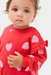 Rød sweatshirt med rosa hjerte-mønstre og flæse detaljer på ærmerne. Blødt stof med rund hals og knapdetalje på skulderen.