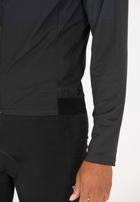 Schwarze Langarmjacke aus strukturiertem Stoff, mit einem Reißverschluss und tailliertem Schnitt. Kombiniert mit schwarzen Radhosen.