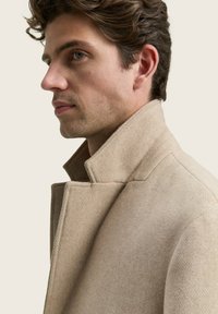 Perfil de un hombre con cabello corto y rizado, vistiendo un abrigo de lana beige con el cuello levantado, frente a un fondo sencillo.
