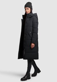 Abrigo de plumas negro con capucha, con secciones acolchadas, bolsillos laterales y cierres a presión. Combinado con pantalones ajustados negros y botines.