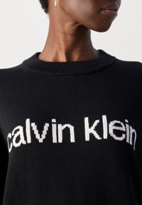 Personne portant un pull en maille noir avec le texte blanc "Calvin Klein", montrant l'encolure et un visage partiel tourné de côté.