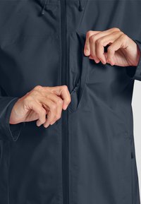 Veste légère bleu foncé avec une fermeture éclair, mettant en avant un gros plan de mains ouvrant une poche latérale. Texture de tissu lisse et durable.
