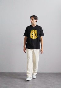 Black t-shirt with a central gold graphic design, paired with cream-colored trousers and white sneakers, standing on a gray floor.