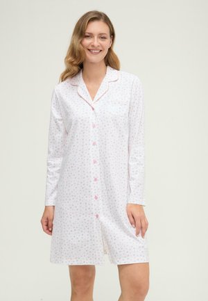 Mujer sonriente con una camisa de dormir blanca de manga larga abotonada con pequeños estampados florales rosas y grises, de pie frente a un fondo liso.