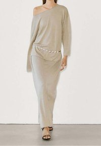 Frau, die in einem beigen, schulterfreien, langärmeligen Kleid mit dekorativem Gürtel und schwarzen offenen Sandalen geht.