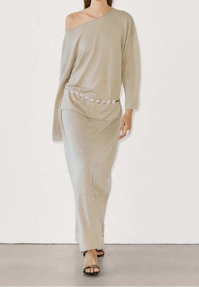 Frau, die in einem beigen, schulterfreien, langärmeligen Kleid mit dekorativem Gürtel und schwarzen offenen Sandalen geht.