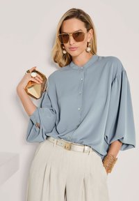 Frau in einer lockeren blauen Knopf-bluse und beigefarbenen Faltenhosen, die eine goldene geometrische Clutch hält, accessorized mit Sonnenbrille und Goldschmuck.
