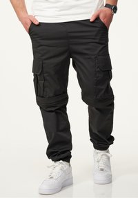 Schwarze Cargo-Hosen mit mehreren Seitentaschen, elastischen Bündchen und einer glatten Textur. Getragen mit weißen Sneakers.