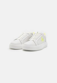 White low-top sneakers with laces, neon yellow details, and "CK Calvin Klein Jeans" logo on the side, photographed on a white background.