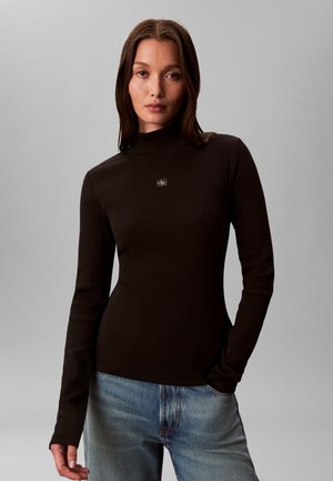 Nainen, jolla on suorat ruskeat hiukset, musta pitkähihaisen poolopaidan yläosa ja siniset farkut, seisoo yksivärisen harmaan taustan edessä.