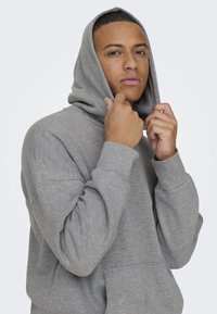 Grauer Kapuzenpullover mit einer Fronttasche, einer Kordelzugkapuze und gerippten Bündchen. Das Material wirkt weich mit einer subtilen Textur.