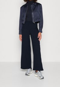 Mörkblå kort satinjacka, svart tröja med hög krage, lösa mörkblå vida byxor och silvriga löparskor med svarta detaljer.