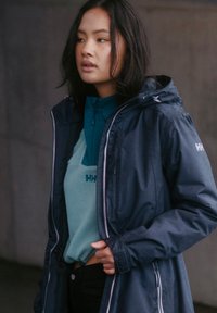 Chaqueta impermeable azul marino con un collar alto y capucha, superpuesta a una camiseta de manga larga verde azulado. Jeans negros, con las manos parcialmente metidas en la chaqueta.