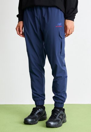 Mężczyzna ubrany w granatowe spodnie dresowe cargo New Balance oraz czarne buty sportowe stoi na jasnozielonej podłodze.