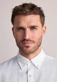 Chemise blanche en lin à boutons visibles, portée par un homme aux cheveux courts et texturés et à la légère barbe, sur un fond neutre.