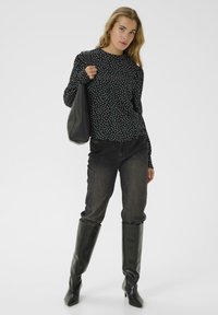 Čierna dlhá rukávová košeľa s bielymi bodkami, spárovaná s tmavými džínami a čiernymi čižmami po kolená. Modelka drží čiernu kabelku cez rameno.