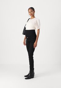 Jeans de maternidad negros con una cintura alta, combinados con una blusa de canalé clara y botas negras de tobillo. Los accesorios incluyen un bolso negro.