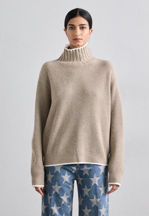 Bež pleteni pulover s turtleneck ovratnikom in belimi detajli, oblečen čez modre jeans hlače z vzorcem zvezd.