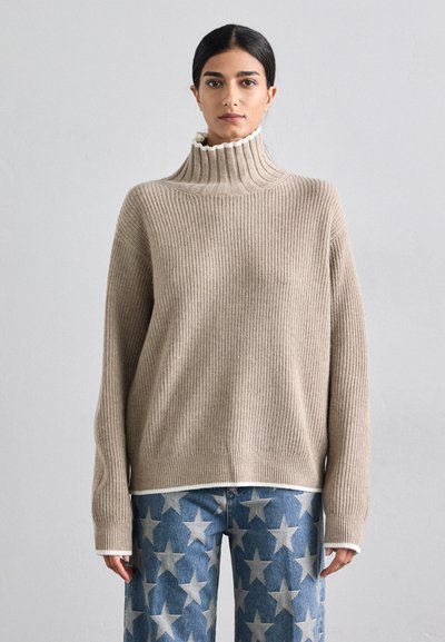 Pull en maille côtelée beige à col montant avec une bordure blanche, porté sur un pantalon en denim bleu à motif étoilé.