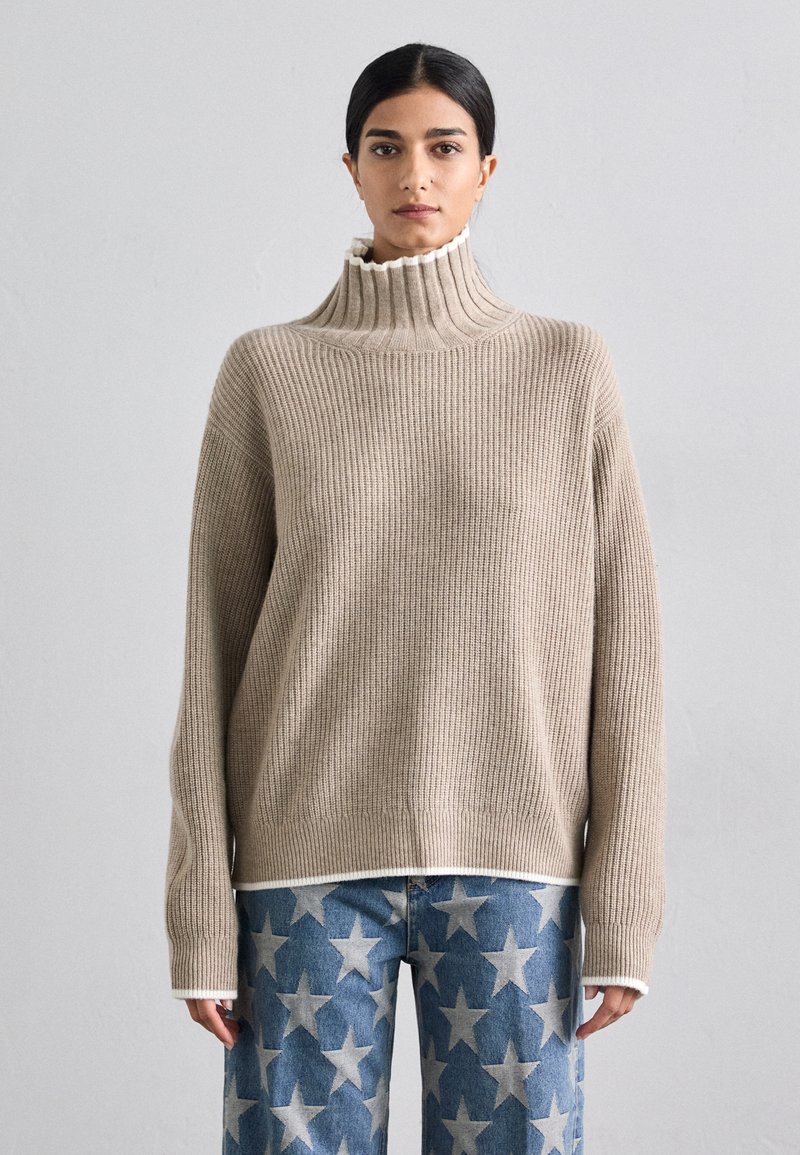 Beige gerippter Rollkragenpullover mit hohem Kragen und weißen Akzenten, getragen über blauen Jeans mit Sternenmuster.