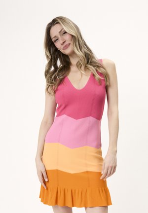 Kvinde med langt blondt hår iført ærmeløs flerfarvet kjole med pink, fersken og orange vandrette striber og flæsekant, smilende.