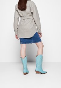 Botas vaqueras azules con puntas puntiagudas, relieve floral y tacón medio apilado, combinadas con una camisa gris con cinturón y una falda vaquera.