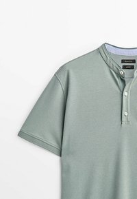 Chemise henley vert sauge à manches courtes avec col bande et trois boutons blancs sur fond uni.