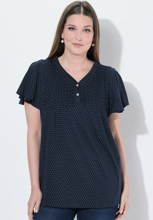 Frau mit glattem braunem Haar, die eine marineblaue, gepunktete Bluse mit kurzen, fließenden Ärmeln und Perlen-Tropfenohrringen trägt.