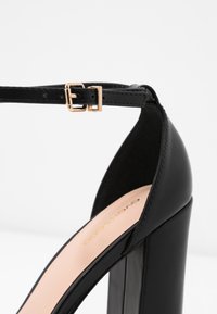 Sandal con tacco alto nero, con una tomaia in pelle lucida, fibbia dorata e un tacco a blocco prominente. Presenta una soletta beige liscia.