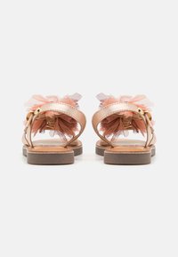 Sandalias metálicas doradas con acentos de tul rosa, suela plana y correa en el tobillo. Diseño de espalda abierta y detalles texturizados en las correas.