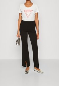 Camiseta blanca con un gráfico triangular rosa, pantalones negros con aberturas laterales y zapatos planos metálicos plateados. Bolso compacto plateado sujetado en la mano.
