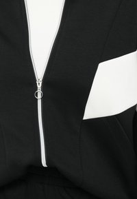 Veste noire avec une fermeture éclair argentée à l'avant et une bande blanche diagonale sur la manche droite, sur fond blanc.