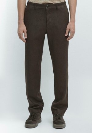 Uomo che indossa pantaloni marrone scuro dritti e scarpe da ginnastica marrone scuro abbinate, in piedi davanti a uno sfondo chiaro e uniforme.