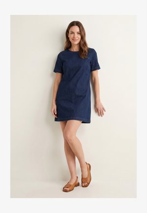 Vrouw met bruin haar tot op de schouders, gekleed in een donkerblauwe korte mouw denimjurk en bruine sandalen met open tenen, staand tegen een witte muur.