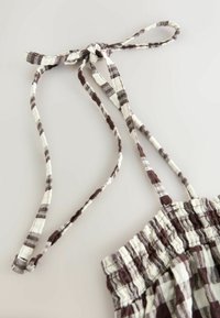 Nœud papillon en tissu à carreaux marron et blanc attaché à une bretelle de vêtement smocké sur un fond clair.
