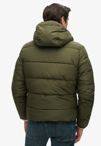 Oliven grøn dunjakke med hætte, der har et quiltet design og elastiske cuffs, båret over blå jeans.