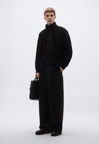 Μαύρο σουέτ μπουφάν με ψηλό γιακά, συνδυασμένο με μαύρο ζιβάγκο, φαρδιά μαύρη παντελόνια και μαύρα δερμάτινα παπούτσια. Κρατά μια μαύρη τσάντα.