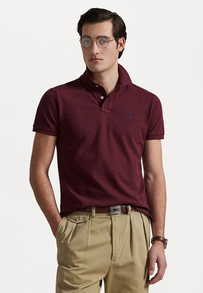 Polo shirt bordeaux avec un col classique, des manches courtes et un petit logo brodé. Porté avec un pantalon beige et une ceinture marron.