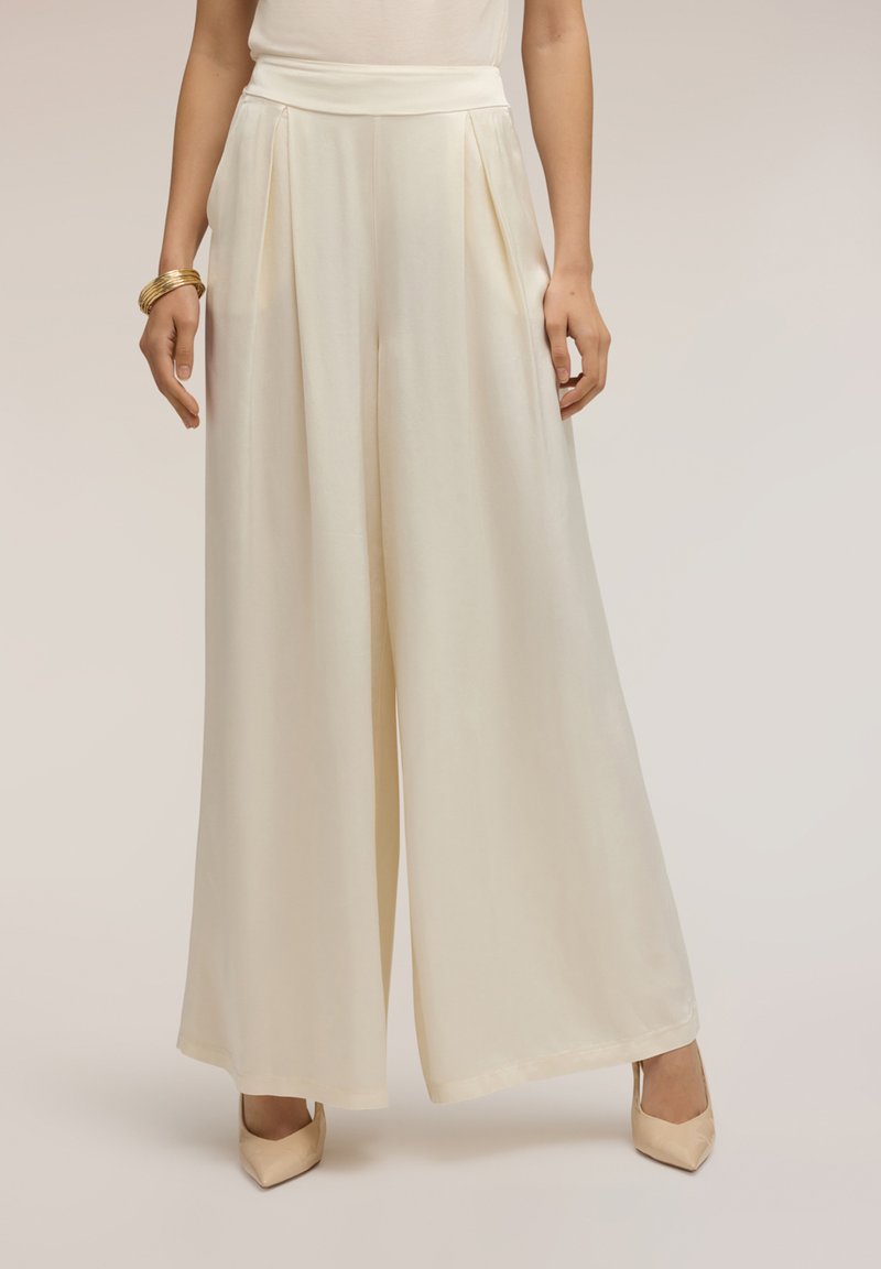 Woman wearing high-waisted, wide-leg cream trousers with pleats, paired with cream pointed-toe heels and gold bangles on her left wrist.