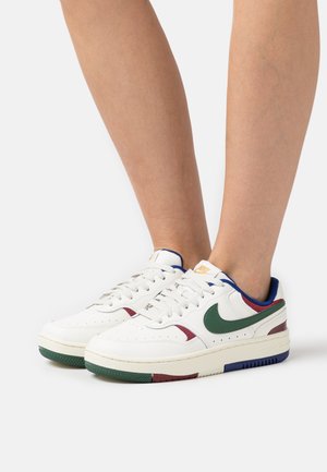 Beine mit weißen Nike-Sneakers mit grünem Swoosh sowie roten und blauen Akzenten, stehend auf weißem Hintergrund.