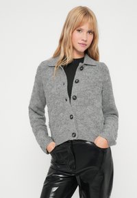 Γκρι πλεκτό γιλέκο με γιακά, κουμπωτή μπροστινή όψη και ριμπ μανίκια. Φοράει από πάνω μία μαύρη μπλούζα, σε συνδυασμό με γυαλιστερά μαύρα παντελόνια.