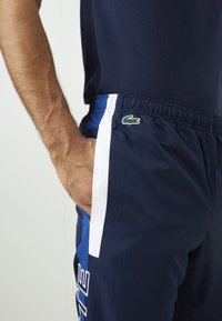 Calções de atletismo azul-marinho com painéis laterais brancos e azuis, cintura elástica e um logótipo verde da Lacoste na coxa esquerda.