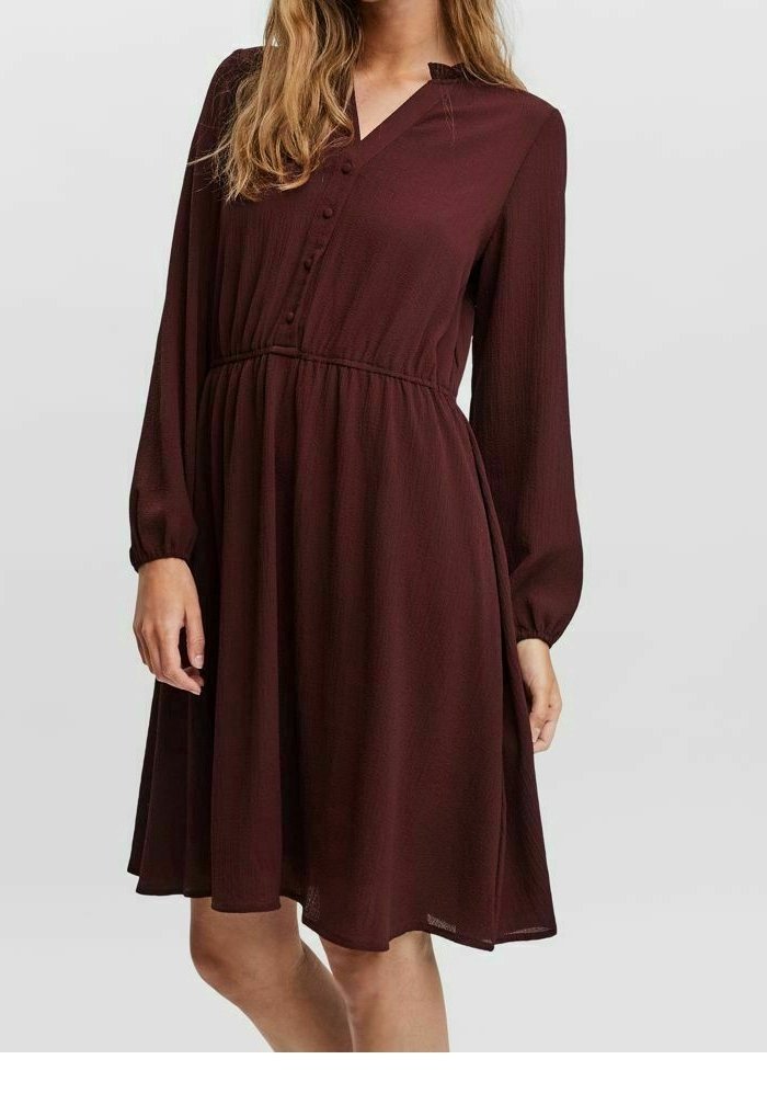 Femme portant une robe bordeaux foncé à manches longues, longueur genou, avec des détails de boutons sur la poitrine et une taille froncée.
