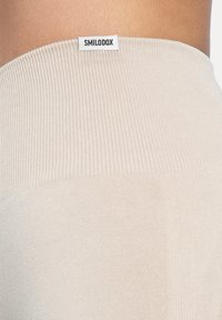 Beżowy prążkowany pas z metką z logo. Tkanina jest gładka o miękkiej fakturze, z dopasowanym krojem, prezentującym czysty i minimalistyczny wygląd.