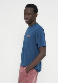 Μπλε βαμβακερό t-shirt με κοντά μανίκια, με πολύχρωμο γραφικό λογότυπο στο στήθος. Φορεμένο με ανοιχτό ροζ παντελόνι κοτλέ.