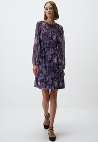 Paarse bloemenjurk met een gerimpelde halslijn, lange doorschijnende mouwen en een getailleerde taille. Het patroon toont grote lichte en donkere bloemen.