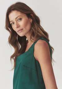 Teal sleeveless top with a round neckline, featuring a small front pocket and a soft, smooth texture. Model displays side profile.