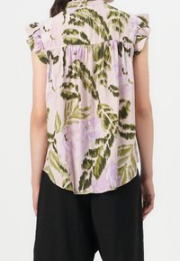 Blouse légère à manches courtes de couleur rose clair avec des motifs floraux verts et violets, accents froncés aux épaules et coupe ample.