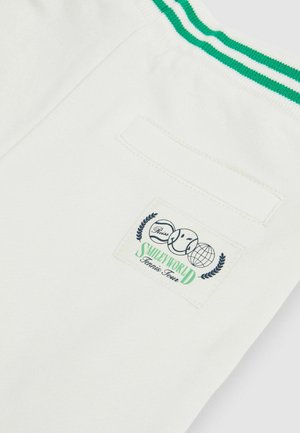 Vita shorts med ett grönt randat midjeband, ett bakfack och en logopatch med ett leende ansikte och en jordglob på tyget.