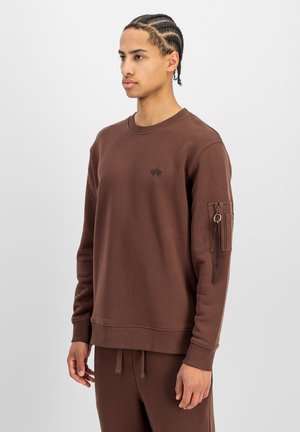Joven con el cabello trenzado que lleva una sudadera marrón con bolsillo con cremallera y detalle de anillo en la manga izquierda, de pie frente a un fondo liso.